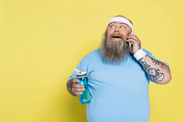 Surpris plus taille homme avec bouteille de sport parler sur téléphone cellulaire isolé sur jaune — Photo de stock