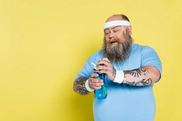 Sediento hombre con sobrepeso con la barba botella deportiva apertura aislado en amarillo - foto de stock
