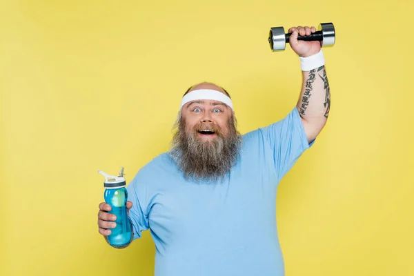 Happy overweight man holding sports bottle while training with dumbbell isolated on yellow — Fotografia de Stock