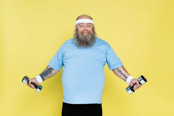 Hombre barbudo alegre con sobrepeso haciendo ejercicio con pesas aisladas en amarillo - foto de stock