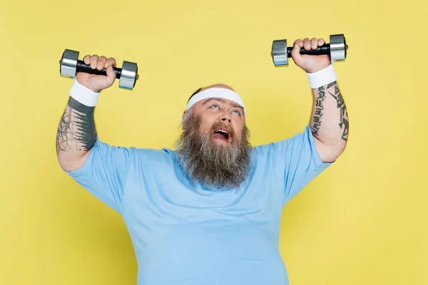 Hombre con sobrepeso haciendo muecas mientras entrenaba con pesadas pesas aisladas en amarillo - foto de stock