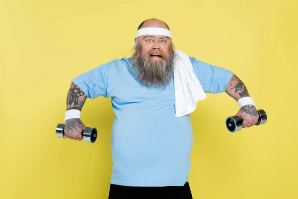 Татуйований надмірна вага людини посміхається під час тренувань з важкими гантелями ізольовані на жовтому — стокове фото