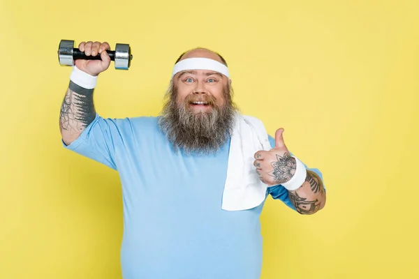 Cheerful plus size man showing thumb up while exercising with dumbbell isolated on yellow — Stock Photo
