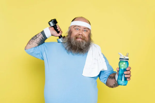 Бородатий плюс розмір чоловік з рушником і спортивною пляшкою вправи з гантелі ізольовані на жовтому — стокове фото