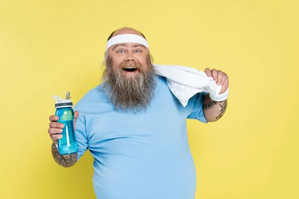 Joyeux homme plus la taille avec bouteille de sport et serviette regardant la caméra isolée sur jaune — Photo de stock