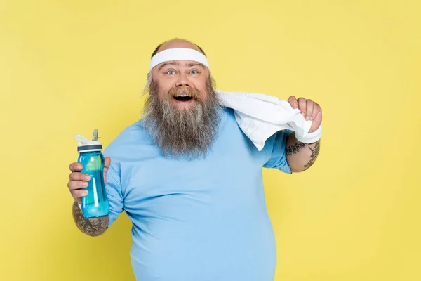 Cheerful plus size man with sports bottle and towel looking at camera isolated on yellow — Stock Photo