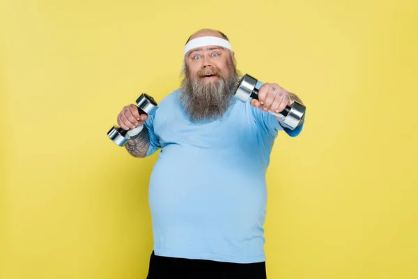 Улыбающийся бородатый мужчина с избыточным весом смотрит в камеру, тренируясь с гантели изолированы на желтый — стоковое фото