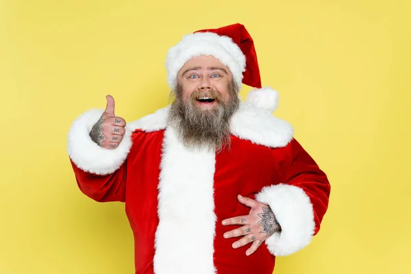 Happy plus size santa claus showing thumb up and touching tummy isolated on yellow — Stock Photo