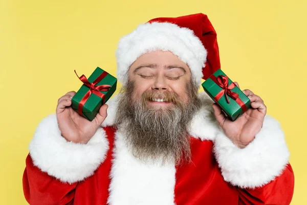 Santa Claus complacido y regordete con los ojos cerrados sosteniendo cajas de regalo aisladas en amarillo - foto de stock