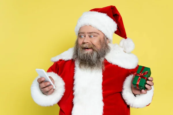 Astonished plus size santa claus with present looking at mobile phone isolated on yellow — Stock Photo