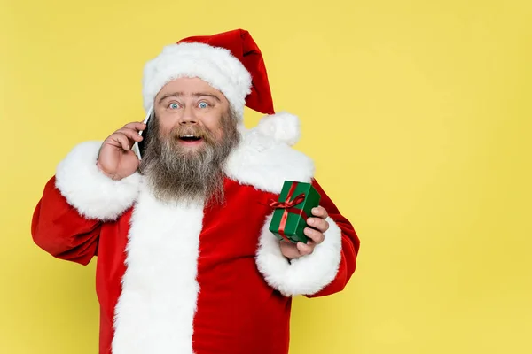 Astonished plus size santa claus showing gift box while talking on smartphone isolated on yellow — Foto stock