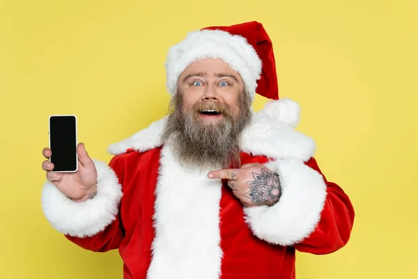 Emocionado sobrepeso santa claus apuntando al teléfono celular con pantalla en blanco aislado en amarillo - foto de stock