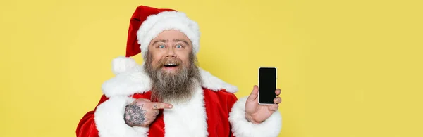 Cheerful plus size santa claus pointing at mobile phone with blank screen isolated on yellow, banner — Foto stock