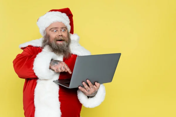 Amazed overweight santa claus pointing at laptop isolated on yellow — Stock Photo