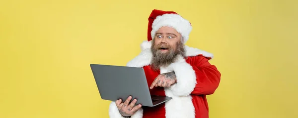 Збуджений плюс розмір чоловіка в різдвяному костюмі вказує на комп'ютер ізольовано на жовтому, банер — стокове фото