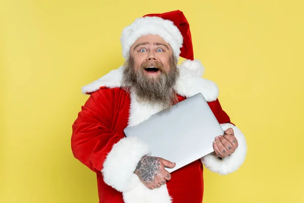 Amazed and happy overweight santa claus holding laptop isolated on yellow — Foto stock
