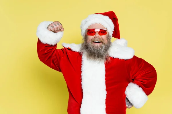 Happy plus size santa claus in trendy sunglasses demonstrating power isolated on yellow — Photo de stock