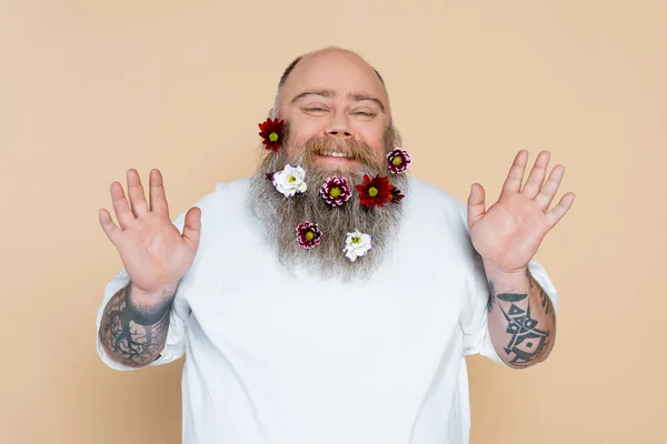 Cheerful plus size man with flowers in beard waving hands at camera isolated on beige — Foto stock