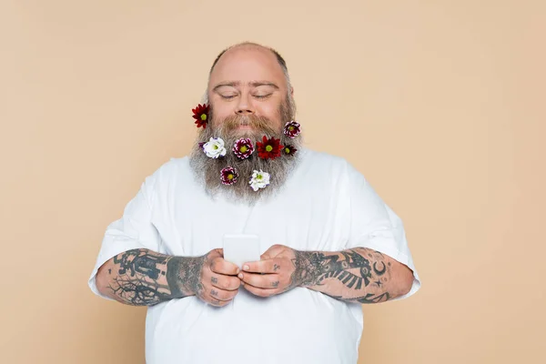 Плюс размер человек с цветочным декором на бороде с помощью смартфона и улыбка изолированы на бежевый — стоковое фото