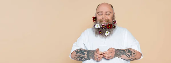 Souriant homme en surpoids avec des fleurs dans la barbe en utilisant un téléphone cellulaire isolé sur beige, bannière — Photo de stock