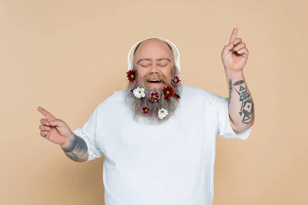 Chubby and cheerful man with decorated beard listening music in headphones isolated on beige — Stockfoto