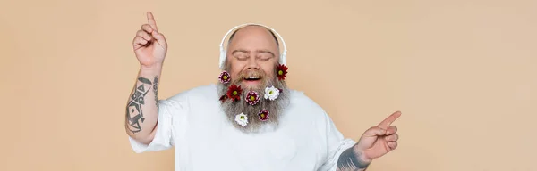 Heureux homme en surpoids avec des fleurs dans la barbe écouter de la musique dans les écouteurs isolés sur beige, bannière — Photo de stock