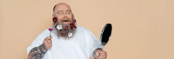 Impressed overweight man with floral decor in beard looking in mirror isolated on beige, banner — Stock Photo