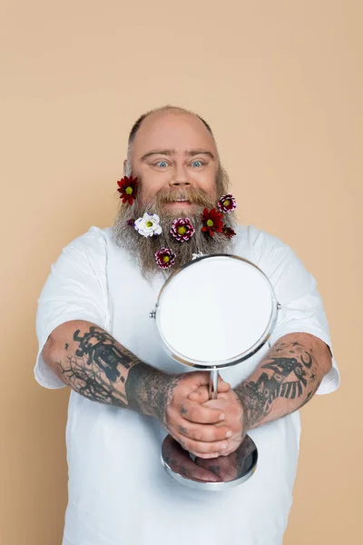 Satisfeito plus size homem com decoração floral em barba olhando no espelho isolado no bege — Fotografia de Stock