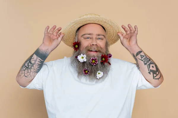 Tattooed overweight man with decorated beard adjusting straw hat and smiling at camera isolated on beige — Stockfoto