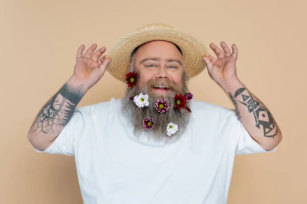 Tatuado excesso de peso homem com barba decorada ajustando chapéu de palha e sorrindo para câmera isolada no bege — Fotografia de Stock