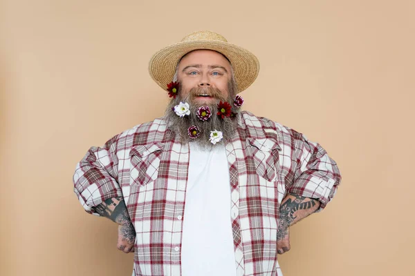 Felice uomo sovrappeso in cappello di paglia e camicia a quadri in posa con fiori in barba isolati su beige — Foto stock