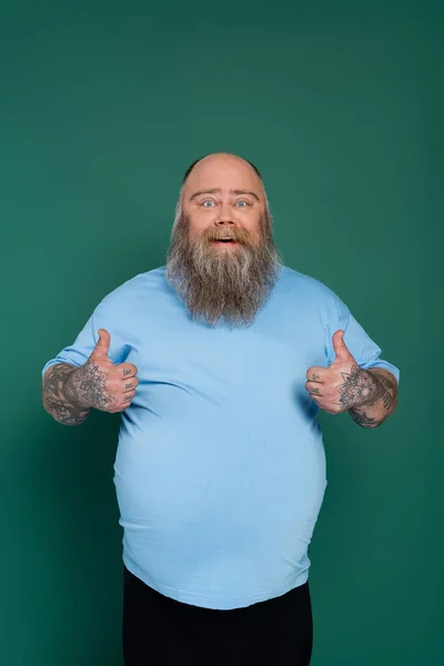 Bearded overweight man in blue t-shirt smiling at camera and showing thumbs up isolated on green — Stock Photo