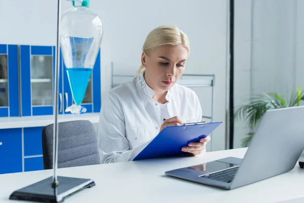 Scientist writing on clipboard near laptop and flask with liquid in lab — Photo de stock
