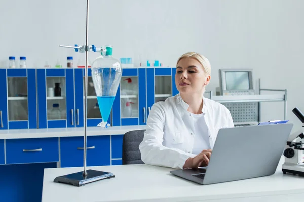 Scientist using laptop and looking at flask with liquid in laboratory - foto de stock