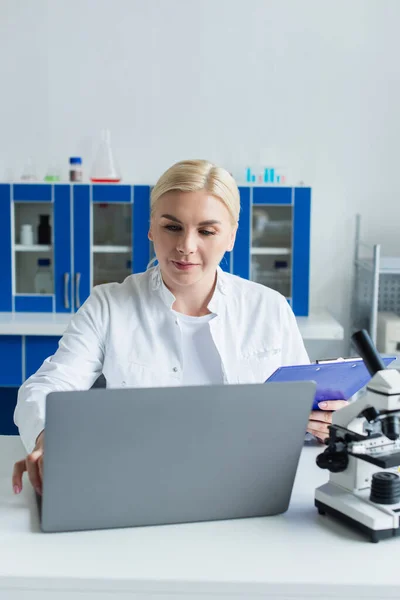 Cientista com área de transferência usando laptop perto de microscópio desfocado em laboratório — Fotografia de Stock