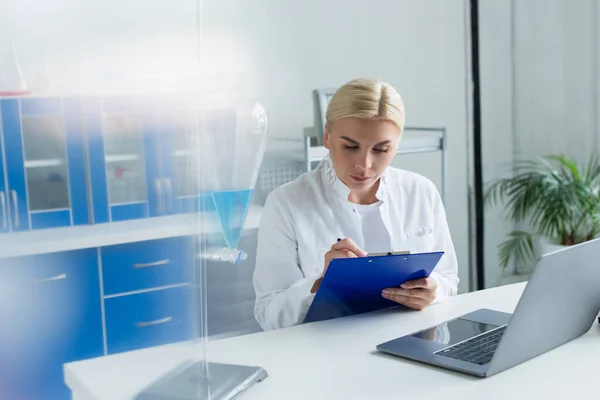 Scientist writing on clipboard near laptop and flask in lab — Stock Photo
