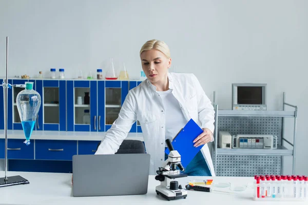 Scientist holding clipboard near laptop and test tubes in lab — стоковое фото