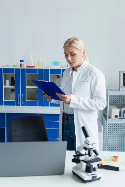 Wissenschaftler hält Klemmbrett in der Nähe von Laptop und Mikroskop im Labor — Stockfoto