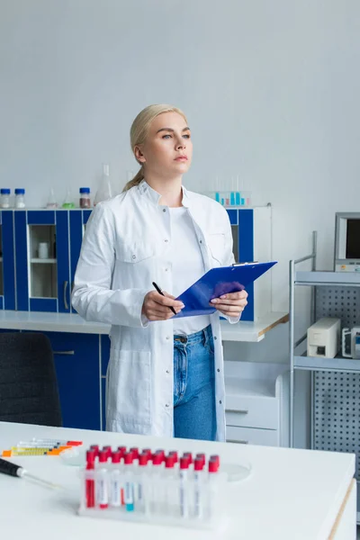 Scientist in white coat holding clipboard near test tubes and electronic pipette in lab - foto de stock