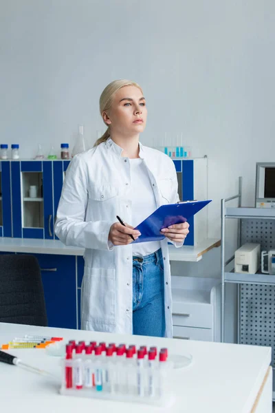 Scientist in white coat holding clipboard near test tubes and electronic pipette in lab — Stock Photo