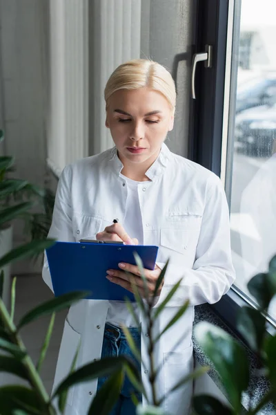 Scientist writing on clipboard near window and plant in lab — Stockfoto
