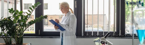 Side view of scientist writing on clipboard near plants and blurred flask in lab, banner - foto de stock