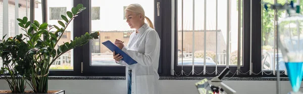 Seitenansicht des Wissenschaftlers beim Schreiben auf Klemmbrett in der Nähe von Pflanzen und verschwommenem Kolben im Labor, Banner — Stockfoto