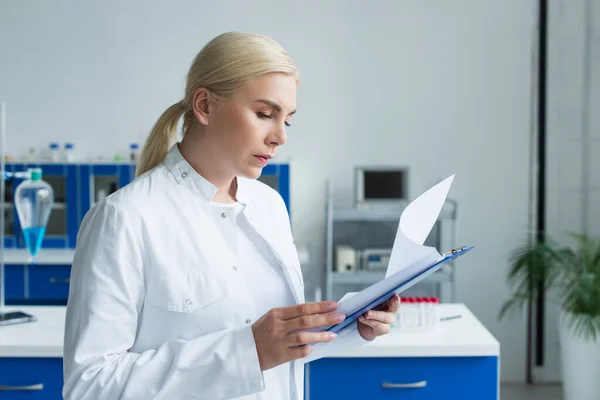 Blonde scientist looking at clipboard while working in lab — Stock Photo