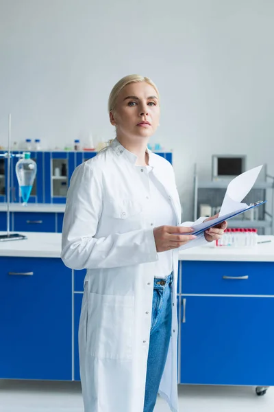 Scientist in white coat holding clipboard and looking at camera in lab — Stock Photo