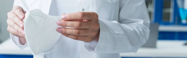 Cropped view of scientist in white coat holding medical mask in laboratory, banner — Stockfoto