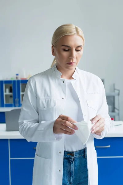 Блондинка-вчений в білому пальто тримає захисну маску в лабораторії — стокове фото