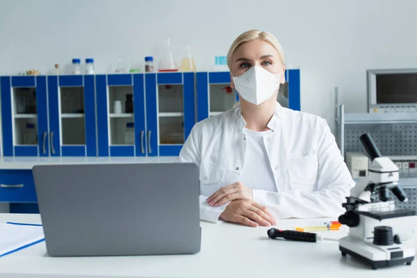 Scientist in protective mask looking at camera near microscope and laptop in lab — Fotografia de Stock