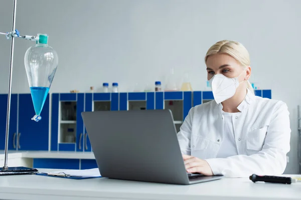 Scientist in protective mask using laptop near flask in lab — Stockfoto