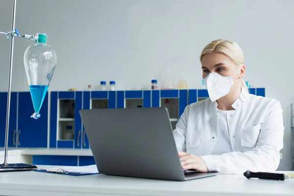 Scientist in protective mask using laptop near flask in lab — Stock Photo